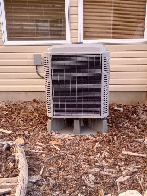 Air conditioning unit outside a house, positioned on a bed of mulch.