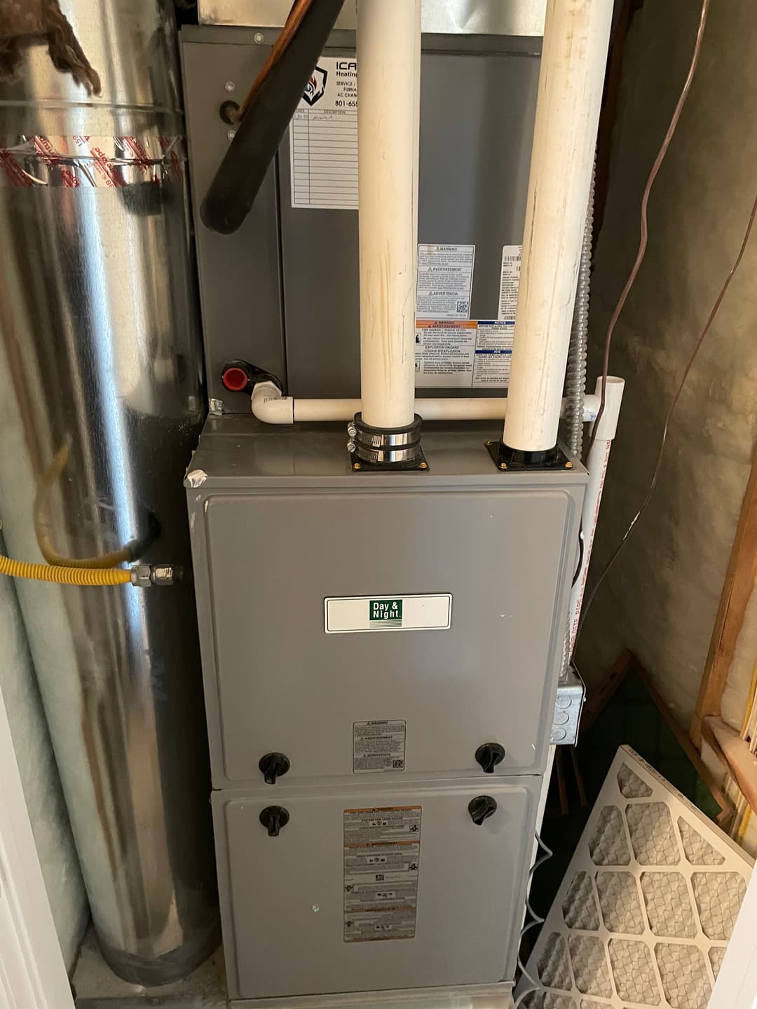 Grey furnace with white pipes, located in a basement corner, near an air filter.