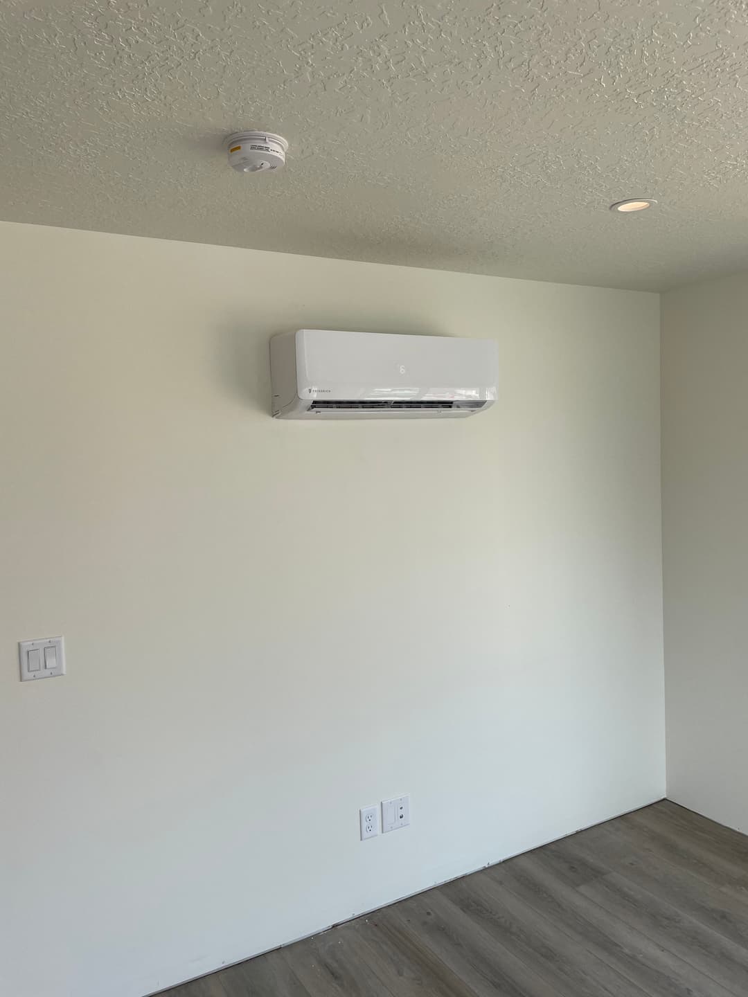 Wall-mounted air conditioner installed in a minimalist room with smoke detector overhead.