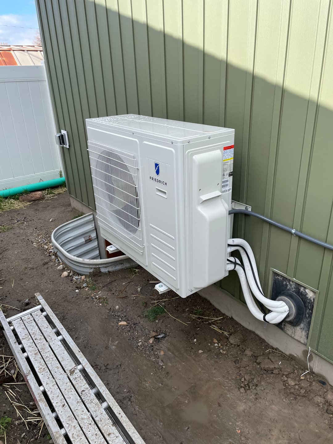 Mini-split air conditioning unit mounted on green exterior wall with drainage piping.