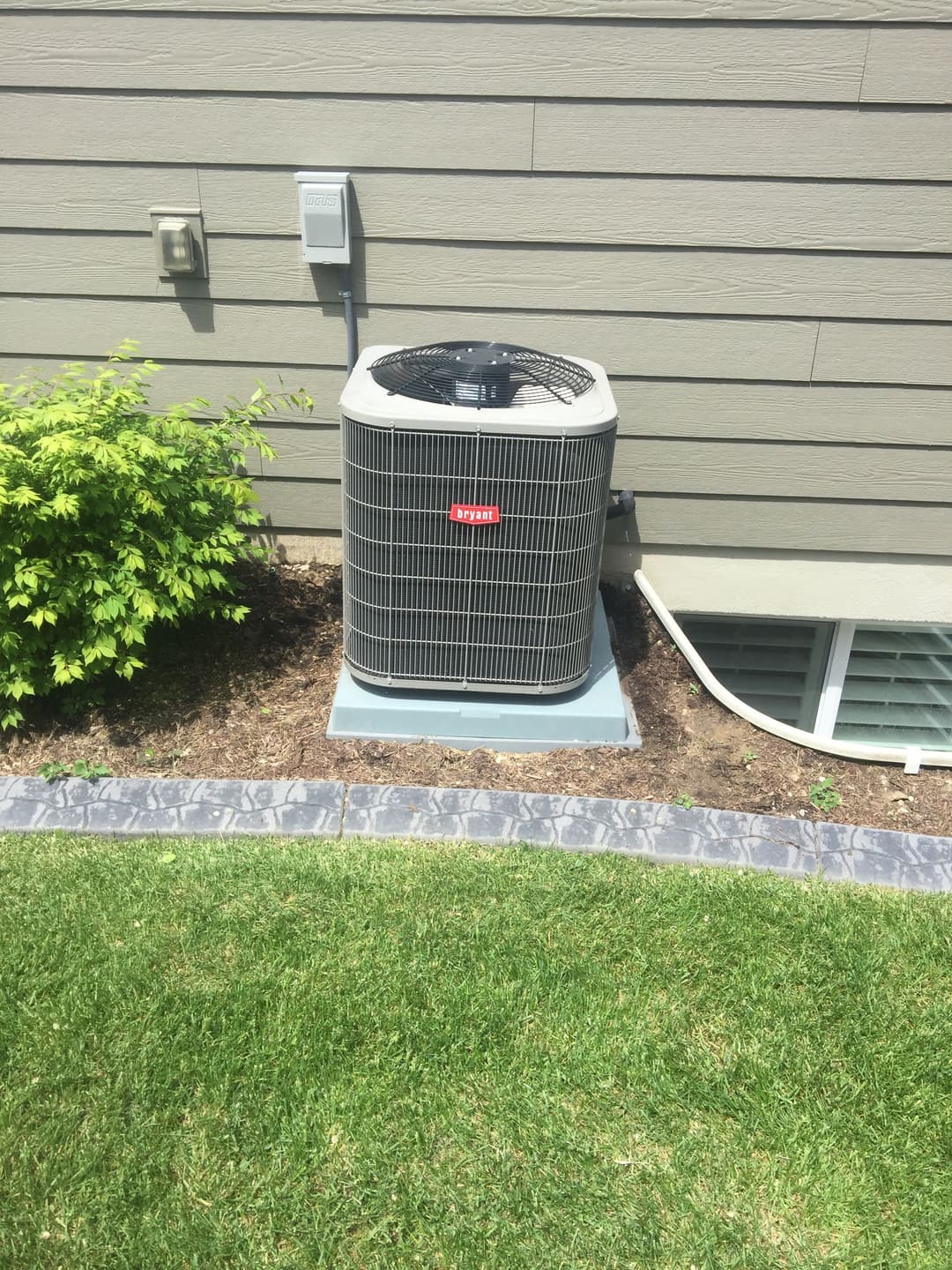 Air conditioning unit installed beside a house, surrounded by landscaped grass and plants.