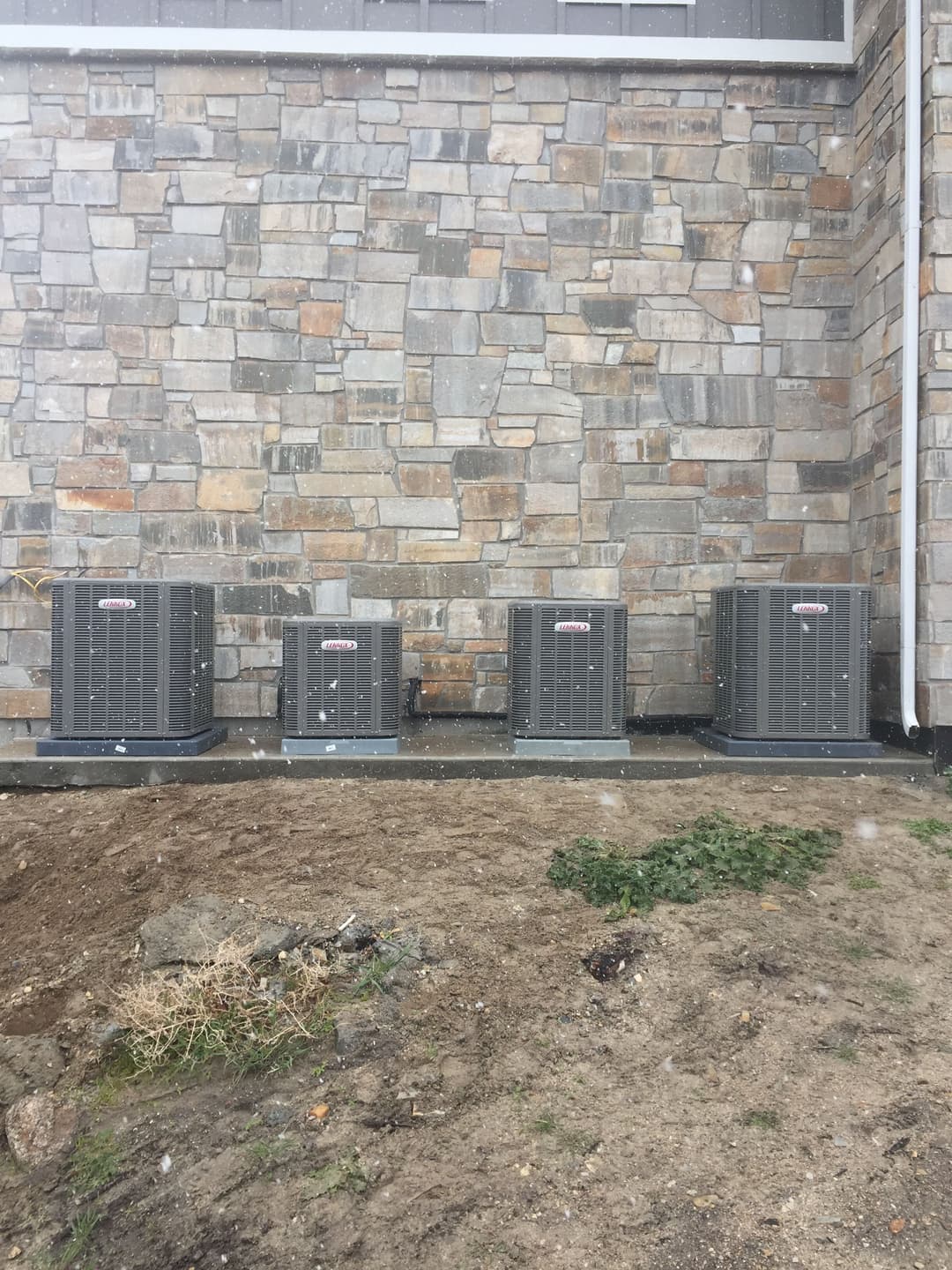 Row of four outdoor HVAC units next to a stone wall in snowy weather.